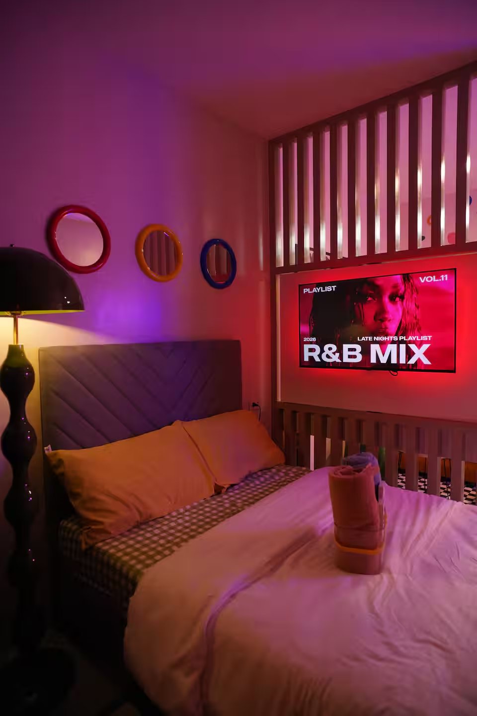 Bedroom in pink ambient glow with R&B Mix on TV, gingham pillow and folded towels on small double bed